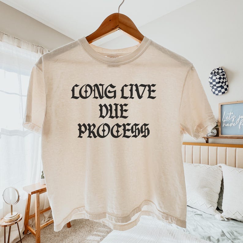 Puede incluir: Camiseta color crema con la frase "LONG LIVE DUE PROCESS" impresa en letras g&oacute;ticas negras. La camiseta est&aacute; colgada en una percha de madera sobre un fondo neutro. Es una prenda informal.