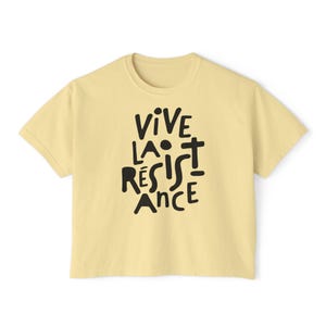 May include: A light yellow t-shirt with the phrase "VIVE LA R&Eacute;SISTANCE" in black, handwritten-style lettering. The shirt has a crew neck and short sleeves. The text is centered on the front of the shirt.
