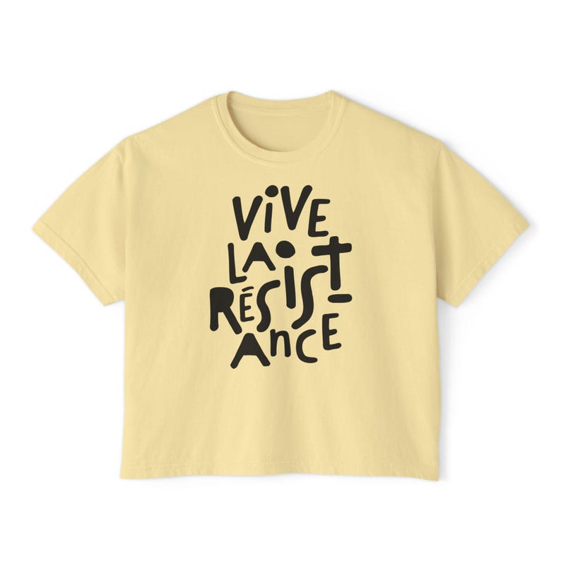 May include: A light yellow t-shirt with the phrase "VIVE LA R&Eacute;SISTANCE" in black, handwritten-style lettering. The shirt has a crew neck and short sleeves. The text is centered on the front of the shirt.