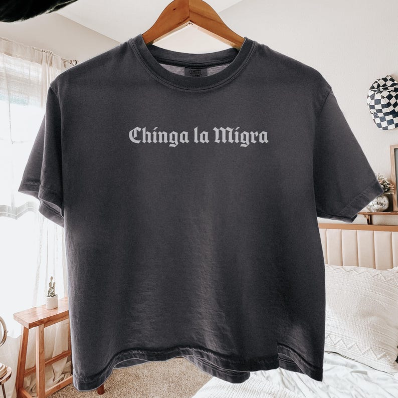 Pode incluir: T-shirt cinzenta escura com a frase "Chinga la Migra" impressa em branco, exibida num cabide de madeira. A camisa tem gola redonda e mangas curtas. O fundo inclui uma cama, uma pequena mesa de madeira e uma janela com cortina.