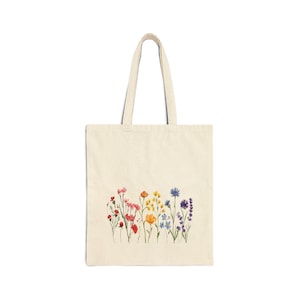 May include: A white canvas tote bag with a colorful floral design. The flowers are arranged in a rainbow pattern, with red, orange, yellow, green, blue, and purple flowers.