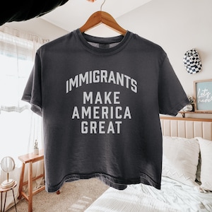 以下が含まれることがあります： 「IMMIGRANTS MAKE AMERICA GREAT」の白文字がプリントされた濃いグレーのTシャツ。木製のハンガーに掛けられています。柔らかく快適な素材で作られたシャツです。