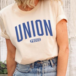 Könnte beinhalten: Ein cremefarbenes T-Shirt mit einer blauen Grafik, auf der in großer Schrift "UNION" und darunter in kleinerer Schrift "PROUD" steht.