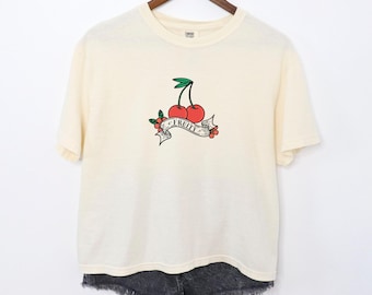 Op de afbeelding: Een crèmekleurig T-shirt met een afbeelding van twee rode kersen met groene stelen en bladeren. De kersen bevinden zich op een banner met de tekst "FRUITY".