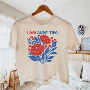 May include: Cream-colored t-shirt with the text "I AM AUNT TIFA" in red and blue. The shirt features a floral design with red flowers, blue leaves, and small red accents. The t-shirt is hanging on a wooden hanger.