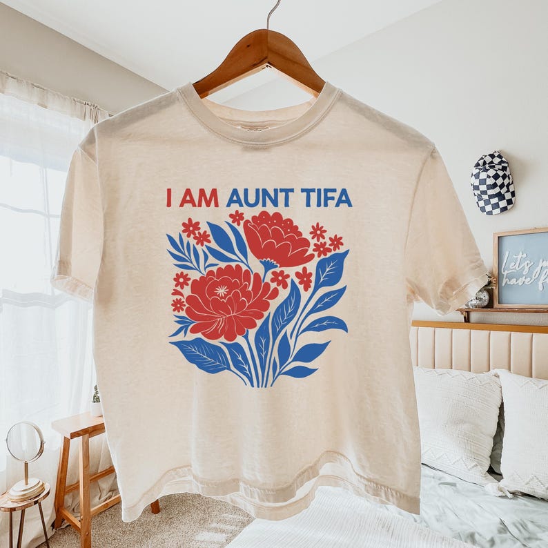 May include: Cream-colored t-shirt with the text "I AM AUNT TIFA" in red and blue. The shirt features a floral design with red flowers, blue leaves, and small red accents. The t-shirt is hanging on a wooden hanger.