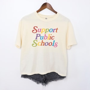 May include: A cream colored t-shirt with short sleeves and a crew neck. The shirt has a graphic print that says "Support Public Schools" in rainbow colors.
