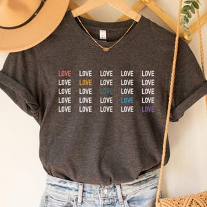 Może przedstawiać: Szary melanżowy t-shirt z okrągłym dekoltem i krótkimi rękawami. Na koszulce wielokrotnie powtarza się słowo "LOVE" w różnych kolorach.