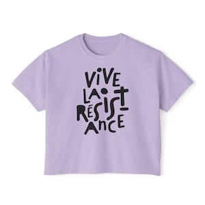 May include: A light purple t-shirt with the words "VIVE LA R&Eacute;SISTANCE" printed in black, stylized lettering. The shirt has a crew neck and short sleeves. The text is centered on the front of the shirt.
