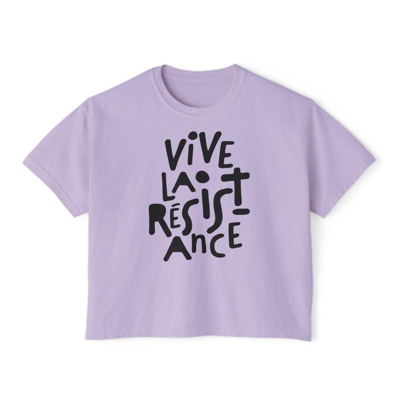 May include: A light purple t-shirt with the words "VIVE LA R&Eacute;SISTANCE" printed in black, stylized lettering. The shirt has a crew neck and short sleeves. The text is centered on the front of the shirt.