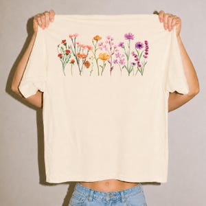 Peut inclure: Un t-shirt à manches courtes de couleur crème orné d'une illustration horizontale à l'aquarelle de fleurs sauvages dans des tons orange, rose et violet sur la poitrine. Le motif représente une rangée de tiges délicates, de boutons et de fleurs.