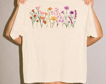 May include: A cream-colored short-sleeve t-shirt featuring a horizontal watercolor illustration of various wildflowers in shades of orange, pink, and purple across the chest. The design depicts a row of delicate stems, buds, and blooming flowers.