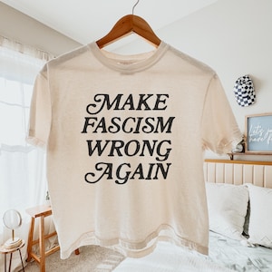 Könnte beinhalten: Cremefarbenes T-Shirt mit dem schwarzen Text "MAKE FASCISM WRONG AGAIN" auf einem Holzbügel. Das Shirt hat kurze Ärmel und einen Rundhalsausschnitt. Der Hintergrund umfasst ein Bett, einen kleinen Tisch und dekorative Gegenstände.