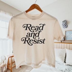 May include: Cream-colored t-shirt with the words "Read and Resist" in a black, decorative font. The shirt is hanging on a wooden hanger. The overall aesthetic is casual and comfortable.