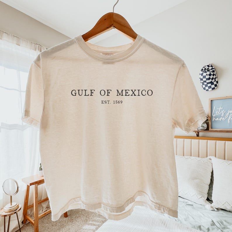 Op de afbeelding: Een cr&egrave;mekleurig T-shirt met de tekst "GULF OF MEXICO EST. 1569" in donkerbruine letters. Het shirt hangt aan een houten hanger. De achtergrond omvat een bed, een kleine tafel en een decoratieve hoed.