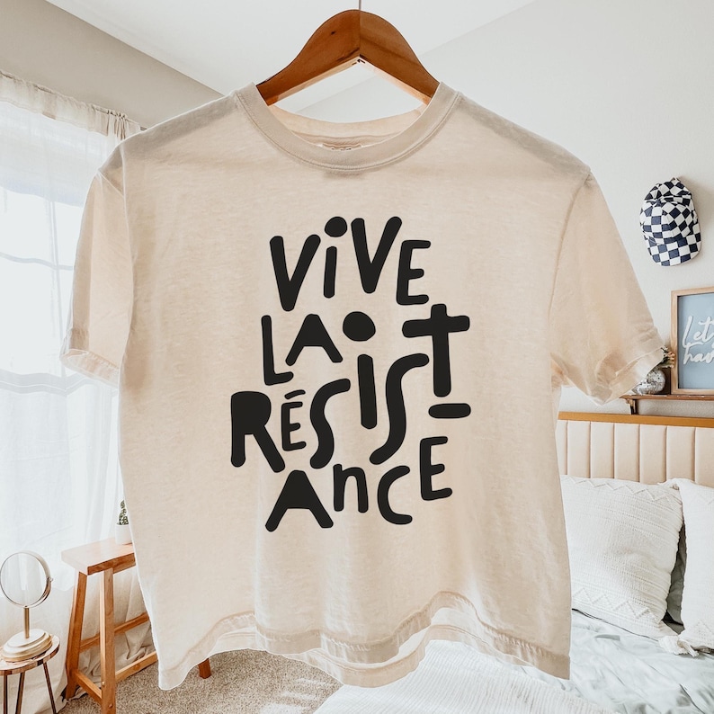 May include: Cream-colored t-shirt with the words "VIVE LA R&Eacute;SISTANCE" in bold black letters. The shirt is hanging on a wooden hanger. The text is in a stylized, artistic font.