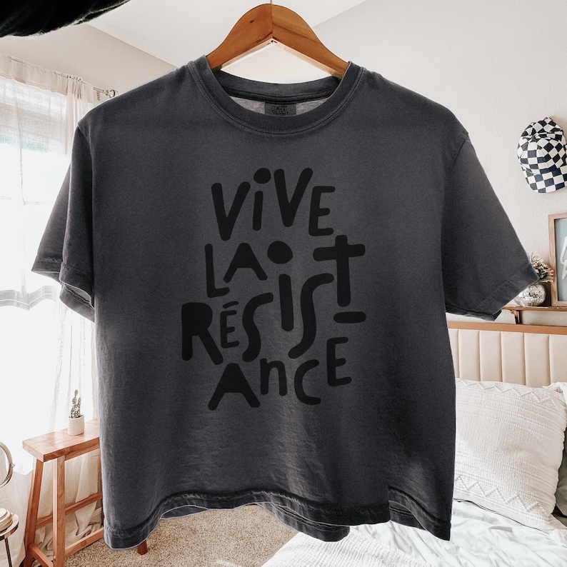 May include: Dark gray t-shirt with the words "VIVE LA R&Eacute;SISTANCE" in black block letters. The shirt is hanging on a wooden hanger. The t-shirt has a crew neck and short sleeves.