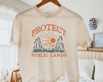 May include: Cream-colored t-shirt with the words "PROTECT PUBLIC LANDS" in orange and blue. The design features a sun, mountains, and cacti. The shirt is hanging on a wooden hanger.