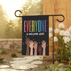 May include: A black garden flag with the text "Everyone is welcome here" in rainbow colors. The flag features a design of multiple hands reaching up with red hearts above them.