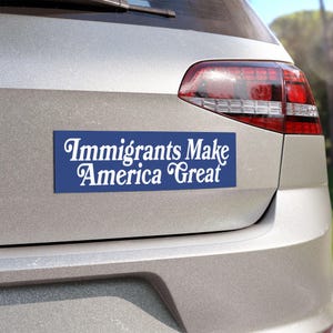 以下が含まれることがあります： 「Immigrants Make America Great」の白い文字が書かれた青い長方形のバンパーステッカー。ステッカーはシルバーの車の後部に貼られています。テキストは筆記体です。