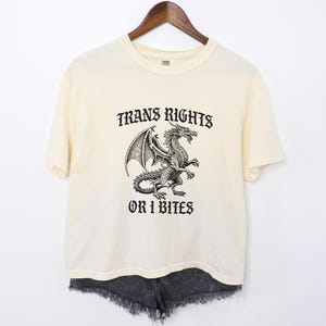 Op de afbeelding: Een crèmekleurig T-shirt met een zwart-witte illustratie van een draak en de tekst "Trans Rights or I Bites".