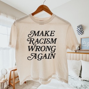 May include: Cream-colored t-shirt with the message "MAKE RACISM WRONG AGAIN" in black script. The shirt is hanging on a wooden hanger. The tee shirt is a casual wear item.