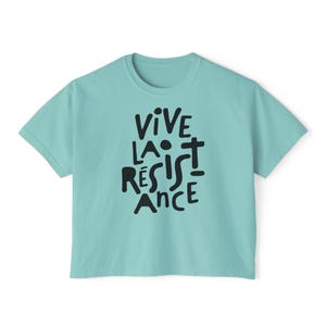 May include: A light teal t-shirt with the words "VIVE LA R&Eacute;SISTANCE" in black, stylized lettering. The shirt has a crew neck and short sleeves. The text is centered on the front of the shirt.
