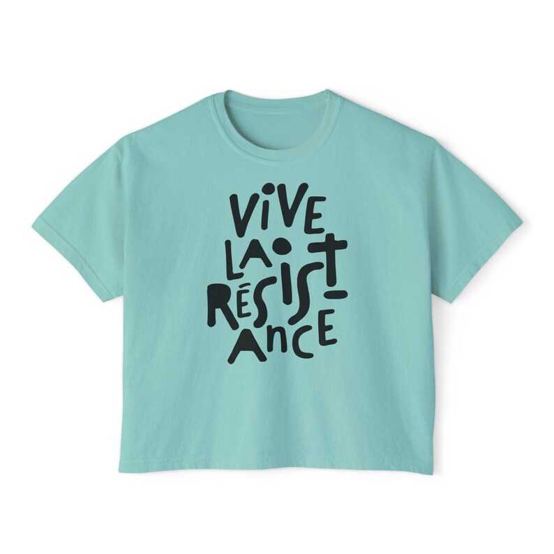 May include: A light teal t-shirt with the words "VIVE LA R&Eacute;SISTANCE" in black, stylized lettering. The shirt has a crew neck and short sleeves. The text is centered on the front of the shirt.