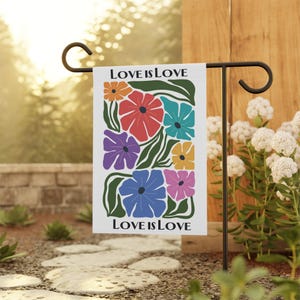 Può includere: Una bandiera da giardino bianca con un motivo floreale e il testo "LOVE IS LOVE". I fiori sono in tonalità di rosso, arancione, giallo, blu, viola e rosa.