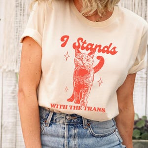 May include: A cream colored t-shirt with a red graphic of a cat with the text "I Stands With The Trans" printed on it.