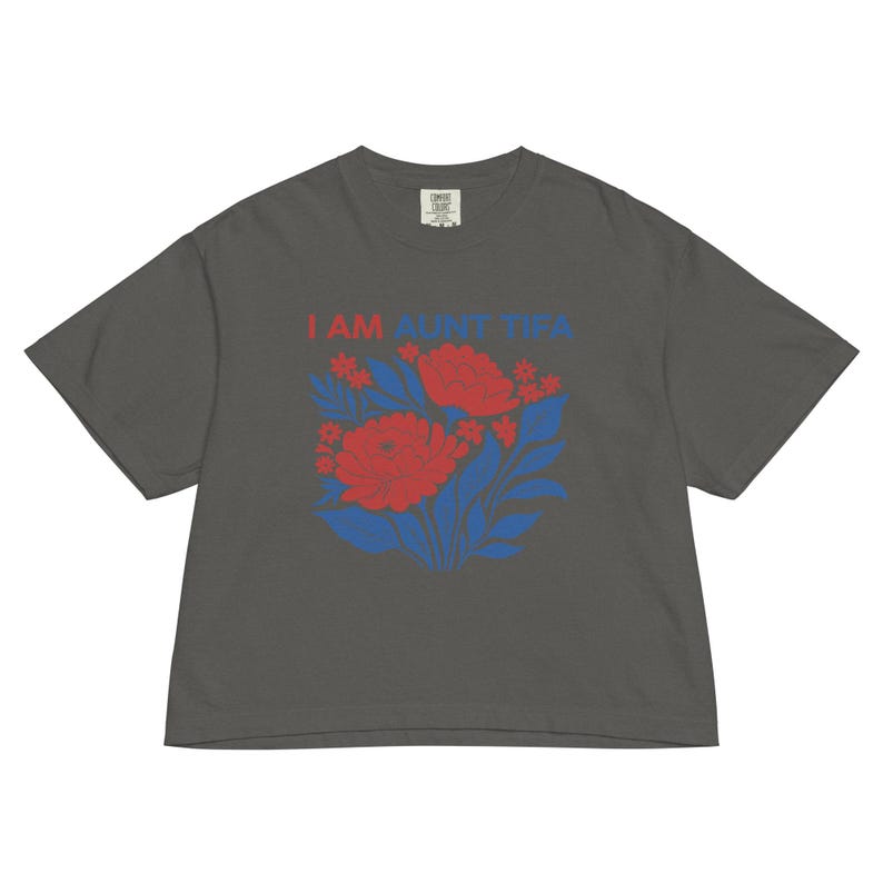 May include: Dark gray t-shirt with the text "I AM AUNT TIPA" in red and blue. The shirt features a floral design with red flowers and blue leaves. The shirt is made of a soft, comfortable material.