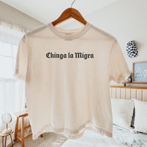 Pode incluir: Uma camiseta creme com a frase "Chinga la Migra" impressa em preto, com letras em estilo g&oacute;tico. A camiseta est&aacute; pendurada em um cabide de madeira.
