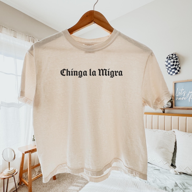 Pode incluir: Uma camiseta creme com a frase "Chinga la Migra" impressa em preto, com letras em estilo g&oacute;tico. A camiseta est&aacute; pendurada em um cabide de madeira.