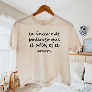 May include: Cream-colored t-shirt with short sleeves hanging on a wooden hanger. The shirt features black text that reads "Lo único más poderoso que el odio, es el amor" with a small red heart. The shirt is made of a soft, lightweight material.
