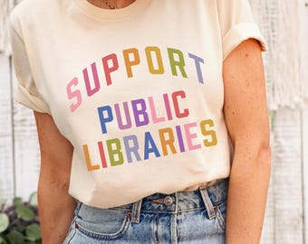 May include: A cream colored t-shirt with the text "SUPPORT PUBLIC LIBRARIES" in rainbow colors.