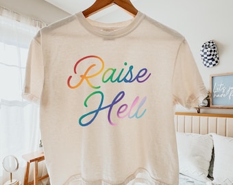 Könnte beinhalten: Ein cremefarbenes Kurzarm-T-Shirt mit dem Schriftzug "Raise Hell" in einer mehrfarbigen Verlaufsschrift. Das T-Shirt hängt an einem Holzkleiderbügel und verfügt über einen Rundhalsausschnitt sowie einen geraden Saumverlauf.