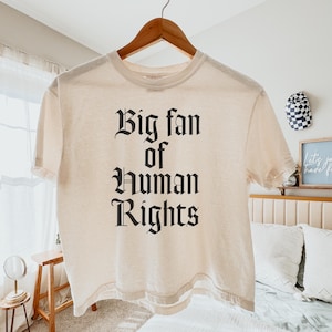 Op de afbeelding: Een crèmekleurig T-shirt met de tekst "Big fan of Human Rights" in een gotisch lettertype. Het shirt hangt aan een houten hanger tegen een neutrale achtergrond. Het T-shirt heeft korte mouwen en lijkt van een zacht materiaal te zijn.
