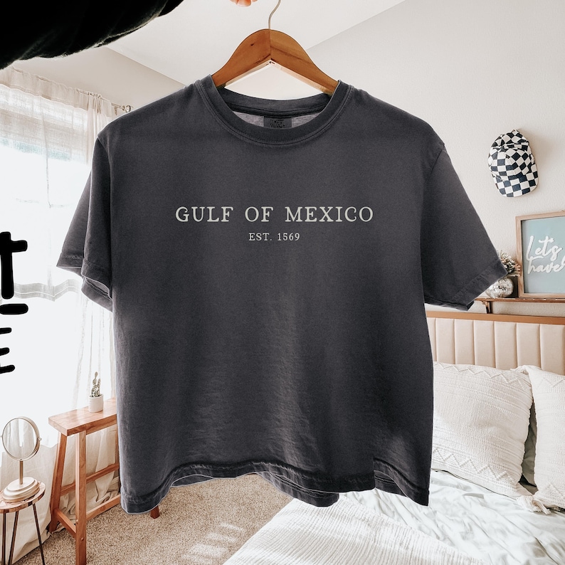 Op de afbeelding: Donkergrijs t-shirt met de witte tekst "GULF OF MEXICO EST. 1569". Het shirt hangt aan een houten hanger. Het t-shirt heeft korte mouwen en een ronde hals.