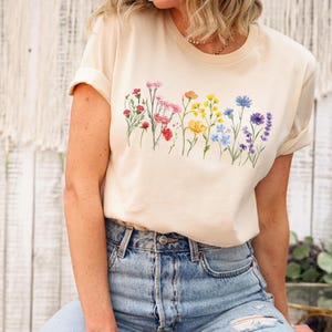 May include: A cream-coloured t-shirt with a watercolour floral design across the chest. The design features a variety of colourful flowers, including red, pink, yellow, and blue. The shirt has short sleeves and a crew neck.