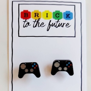 以下が含まれることがあります： カラフルなボタンが付いた黒いビデオゲームコントローラーのイヤリングのペア。イヤリングは、「BRICK to the future」というテキストが書かれた白いカードの上にあります。