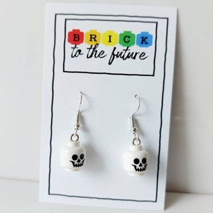 May include: White Lego minifigure earrings with a skull face design. The earrings are silver and have a hook closure. The earrings are displayed on a white card with the text "BRICK to the future".