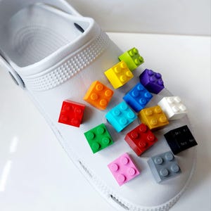 May include: A white clog with colourful Lego blocks attached to the top. The blocks are in various colours, including red, blue, green, yellow, orange, purple, and white.