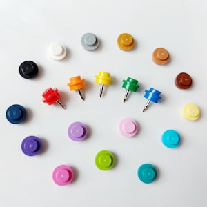 May include: Assortment of colorful plastic push pins in various colors, including red, orange, yellow, green, blue, purple, black, white, brown, and gray. The push pins are arranged in a circle on a white background.