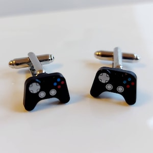 May include: A pair of silver cufflinks with black game controller designs. The controllers have a gray button, a red button, and a blue button.