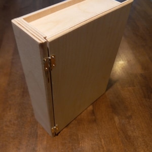 May include: A light-colored wooden box with a hinged lid and brass-colored hardware. The box is rectangular and appears to be made of unfinished wood. The lid has a small lip on the top. The box is sitting on a wooden surface.
