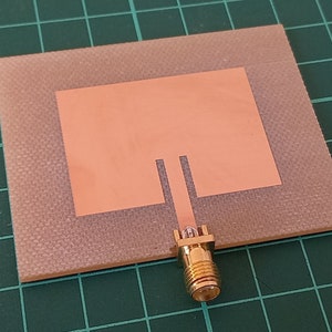 Peut inclure: Une petite antenne carrée de couleur cuivre avec un connecteur doré. L'antenne est montée sur une surface beige texturée. La surface est étiquetée "CNC".