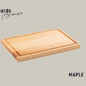 Puede incluir: Una tabla de cortar rectangular de madera de arce con un borde elevado y una ranura alrededor del perímetro. MAPLE está impreso en azul debajo de la tabla.