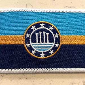 Op de afbeelding: Geborduurde patch met een vlagontwerp. De bovenkant is blauw, met een gouden streep en marineblauwe basis. Een cirkelvormig embleem met witte pilaren en sterren is gecentreerd. De patch heeft een witte rand.