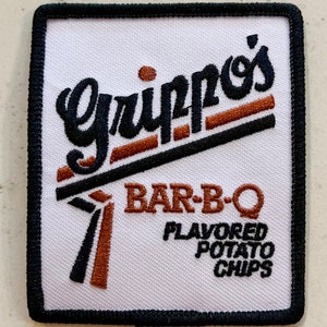 May include: A white embroidered patch with black and brown stitching. The patch features the text "Gripp's" in a stylized font with a brown outline. Below the text is "BAR-B-Q" in brown text and "FLAVORED POTATO CHIPS" in black text.