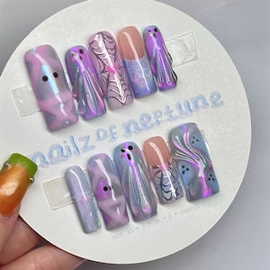 May include: A set of ten press-on nails featuring a pastel purple, pink, and blue color palette with ghost, spiderweb, and star designs. The nails are displayed on a circular card labeled "nailz of neptune" with size options listed below.